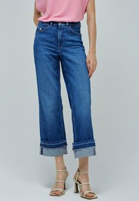 Pantalones vaqueros de mezclilla anchos en azul con dobladillo deshilachado, puños enrollados y diseño clásico de cinco bolsillos. Combinados con una blusa rosa claro y tacones con tiras.