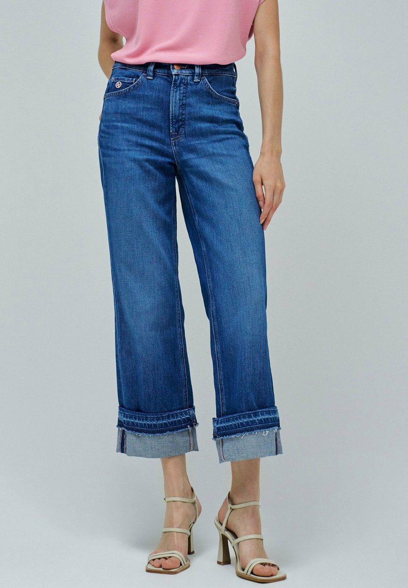 Pantalones vaqueros de mezclilla anchos en azul con dobladillo deshilachado, puños enrollados y diseño clásico de cinco bolsillos. Combinados con una blusa rosa claro y tacones con tiras.