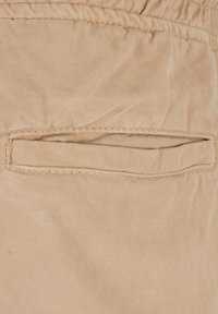 Beiger Stoff mit glatter Textur verfügt auf einer Seite über eine kleine, horizontale Tasche mit einer abgesteppten Nahtdetail.