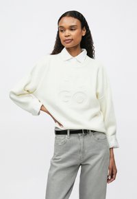 Γυναίκα με λευκό πουλόβερ με ανάγλυφο λογότυπο πάνω από λευκό πουκάμισο και γκρι τζιν, στέκεται με το ένα χέρι στη μέση της μπροστά σε λευκό φόντο.