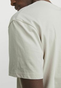 Camiseta de manga corta beige clara con cuello redondo, hecha de suave algodón. Presenta costuras simples en los hombros y en el dobladillo.