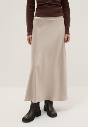 Mujer con falda beige de línea A, blusa de manga larga marrón oscuro y botines negros, posando frente a un fondo claro y sencillo.