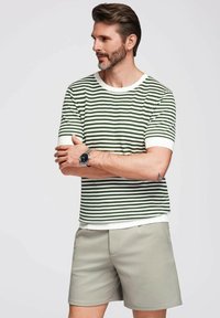 Camiseta de punto a rayas verdes y blancas con cuello redondo, combinada con pantalones cortos ligeros de color beige. La camiseta presenta una textura acanalada y mangas cortas.