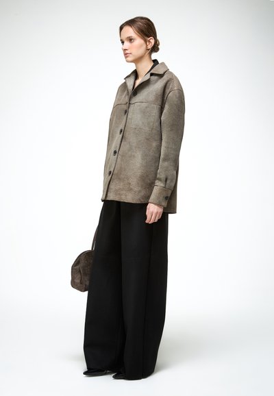 Žena s nízkym účesom v cope, oblečená v taupe zapínanej bunde a širokých čiernych nohaviciach, drží po boku hnedú textúrovanú kabelku.