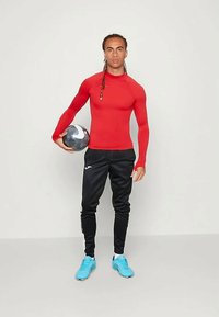 Punainen pitkähihainen urheilupaita yhdistettynä mustiin housuihin, pitäen mustaa ja hopeista palloa. Jalkineet ovat vaaleansiniset jalkapallokengät.