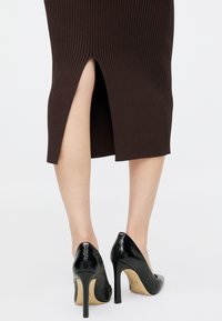 Καφέ φούστα με ραβδώσεις και σχισμή στο πίσω μέρος, συνδυασμένη με γυαλιστερά μαύρα ψηλά τακούνια που διαθέτουν ανάγλυφη υφή και μυτερή γραμμή.