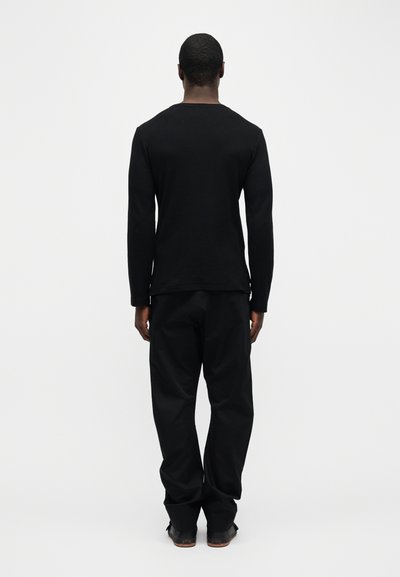 Άνδρας που στέκεται με την πλάτη στραμμένη, φοράει μαύρο μακρυμάνικο πουκάμισο, μαύρο παντελόνι και μαύρα παπούτσια, μπροστά σε ένα απλό φωτεινό φόντο.