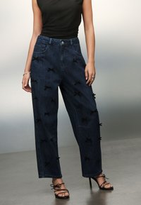 Pantalones de mezclilla de pierna ancha en lavado oscuro con lazos de tela negra en contraste. Cuentan con cierre frontal de botones y bolsillos laterales.