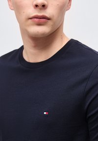 Νεαρός άνδρας που φοράει μπλε σκούρο t-shirt με λαιμόκοψη και ένα μικρό ορθογώνιο λογότυπο κόκκινου, άσπρου και μπλε χρώματος στο στήθος.