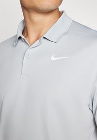 Svetlo siva polo majica z klasičnim ovratnikom, dvema gumboma na zapenjanju in majhnim belim logotipom Nike na prsih. Gladek teksturiran material.