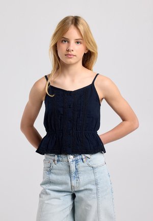 Blond meisje met een zwart mouwloos topje en lichtblauwe hoge taille jeans, staand met handen achter haar rug tegen een grijze achtergrond.