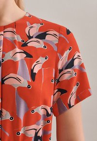 Röd blus med korta ärmar, med ett färgglatt mönster av flamingos i rosa, svart och lavendel. Slät och lätt tygtextur.