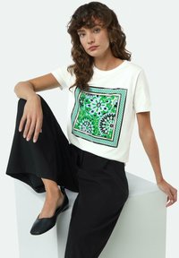 Witte katoenen t-shirt met een groen en blauw bloemenmotief in een vierkant ontwerp. Gepaarde met losse zwarte broeken en zwarte platte schoenen.