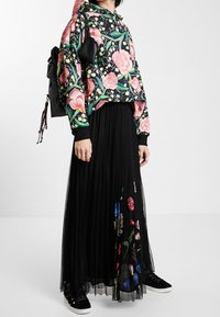 Schwarzer Hoodie mit lebhaften pinken und grünen Blumenmotiven, kombiniert mit einem schwarzen plissierten Rock mit bunten, bestickten Blumen. Schwarze Sneaker.