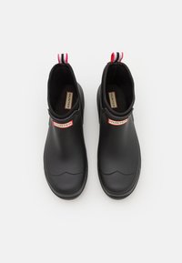 Bottes de cheville en caoutchouc noir avec une texture lisse, présentant un bout arrondi et des tirettes rayées rouges et blanches. Marquées avec "HUNTER" sur les côtés.