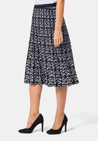 Jupe plissée à motifs noir et blanc avec une taille ajustée, atteignant le milieu du mollet. Associée à des chaussures à talons hauts noires.