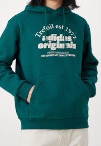 Grön sweatshirt med huva, med en kenguruficka, ribbade manschetter och en vit grafisk logotyp på framsidan. Mjuk textur.