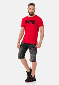 Rotes Baumwoll-T-Shirt mit "WORK"-Aufdruck, kombiniert mit grauen Cargo-Shorts mit Taschen und roten Akzenten, abgerundet mit schwarzen Sportschuhen.
