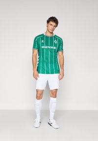 Young man stands wearing a green striped soccer jersey, white shorts, white socks with shin guards, and white cleats against a plain background.