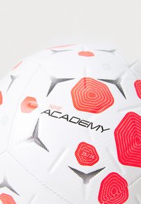 Nike Academy football, white with red and black hexagonal patterns, textured surface, featuring branded logo and number accents.