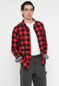 Rot-schwarz kariertes Flanellhemd mit durchgehender Knopfleiste, Brusttasche und denimähnlichen Akzenten an den Manschetten. Kombiniert mit grauer Hose.