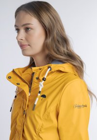 Sárga vízálló dzseki kapucnival, fehér húzózsinórokkal és fekete ernyő logóval a mellrészen. Simább, fényes textúra.