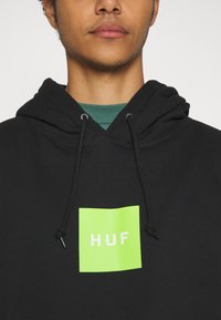 Felpa nera con cappuccio e coulisse. Presenta un logo quadrato verde brillante con la scritta "HUF" in bianco. Il materiale sembra morbido e strutturato.