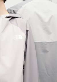 Ανοιχτό γκρι και λεβαντίνια μπουφάν με το λογότυπο "The North Face" στον αριστερό ώμο, φορεμένο από πρόσωπο που βλέπει προς τα πίσω, φόντο ανοιχτό γκρι μπουφάν.