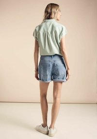 Top corto de manga corta verde claro y texturizado con la espalda plisada, combinado con shorts de mezclilla azul claro y zapatillas blancas.
