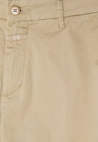 Pantalon beige avec fermeture à bouton, passants de ceinture, poche latérale, et une petite étiquette de la marque en tissu près de la poche.