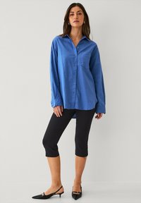 Blauwe, lange mouwen overhemd met puntige kraag en borstzak, gecombineerd met zwarte capri-broeken en puntige zwarte schoenen.