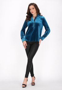 Bluză din voal de mătase teal cu mâneci lungi și guler inalt, având trei nasturi pe plastron. Asortată cu colanți negri elastici și tocuri.