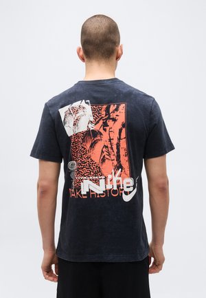 Homme aux cheveux très courts portant un t-shirt noir avec un motif abstrait rouge et blanc et un texte partiel « Nike » dans le dos.