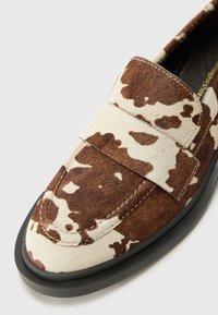 Brown and white cow-patterned loafers made from textured suede, featuring a flat black rubber sole and a wide strap across the vamp.