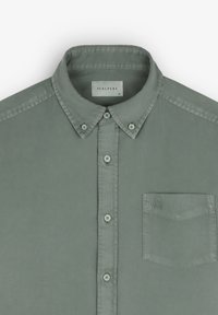 Světlá zelená košile na knoflíky s límečkem, náprsní kapsou a vyšitým logem. Tkanina má hladkou texturu a klasický design.