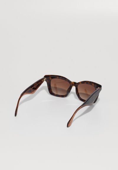 Des lunettes de soleil en écaille de tortue, de forme carrée, avec des verres teintés bruns, posées ouvertes sur une surface blanche.