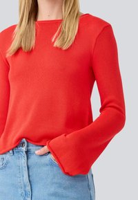 Pull rouge à manches longues avec un tricot texturé et des manches évasées, associé à un jean bleu clair. Caractéristiques : encolure ronde et coupe décontractée.