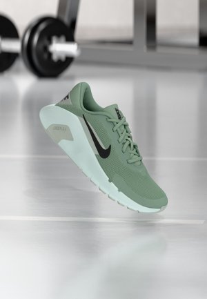 Zaļš Nike sporta apavu pāris ar melnu Swoosh logotipu un baltu zolīti, redzams gaisā virs spoguļojuma treniņu zāles grīdas ar izplūdušiem svaru rīkiem fonā.