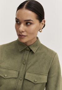 Camicia verde oliva con bottoni, realizzata in tessuto morbido, dotata di due tasche sul petto, colletto e una finitura testurizzata per un tocco di dettaglio in più.