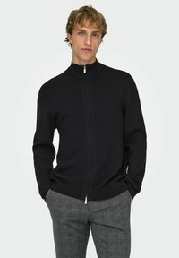 Maglione nero con zip, collo alto, lavorazione a maglia e sottili cuciture verticali. Indossato con pantaloni grigi fantasia.