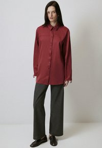 Camicia rossa con bottoni, colletto, maniche lunghe e polsini regolabili; abbinata a pantaloni larghi grigi e scarpe marroni scure.