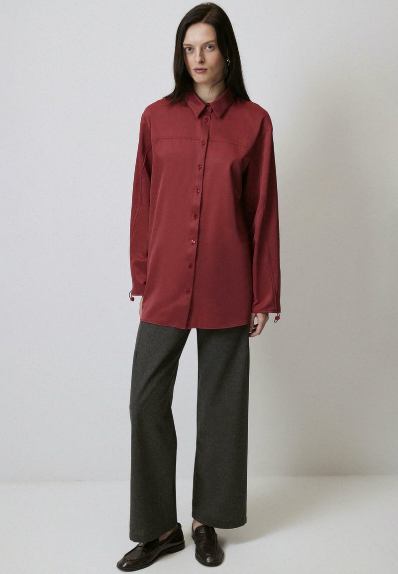Camicia rossa con bottoni, colletto, maniche lunghe e polsini regolabili; abbinata a pantaloni larghi grigi e scarpe marroni scure.