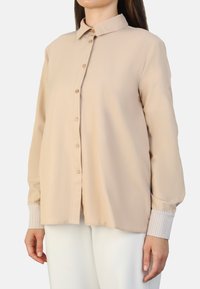 Beige button-up shirt with a classic collar, long sleeves, and striped cuffs. Made from a smooth fabric with a relaxed fit.