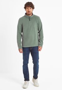 Fleece pullover in muted green with a half-zip collar, textured fabric, and long sleeves, paired with blue jeans and white sneakers.