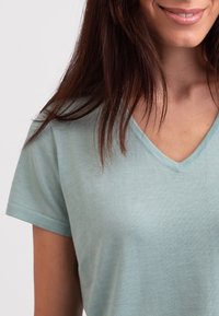 Svetle zelené tričko s výstřihem do V vyrobené z měkké, strukturované tkaniny. Krátké rukávy a volný střih, s jemnými švy podél výstřihu.