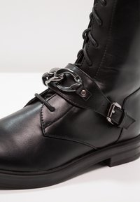 Botte en cuir noir à lacets avec sangle décorative, boucle en argent et détail chaîne à la cheville, dotée d'un petit talon bloc et d'une finition lisse.