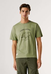 Camiseta de algodón verde oliva con mangas cortas, cuello redondo y gráfico impreso de una furgoneta con el texto "LAS COSAS SUCEDEN, VE UN PASO MÁS ALLÁ."