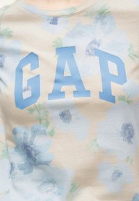 Gros plan d'une chemise beige avec des motifs floraux bleu pâle et vert et de grandes lettres bleues "GAP" sur la poitrine.