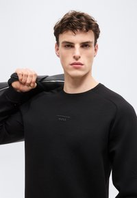 Jeune homme aux cheveux bruns courts portant un sweat-shirt noir avec le logo "Porsche BOSS", tenant une veste noire sur son épaule.