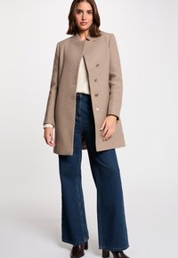 Abrigo de lana beige con un diseño recto, que presenta un cuello mao, seis botones dorados y una textura suave. Combinado con jeans de denim de pierna ancha.